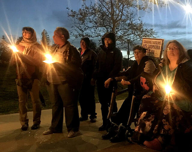 Photo of 6 cohort members at a vigil resisting Islamophobia. 3 are holding burning candles; 4 are standing; 2 are in wheelchairs; 1 has a sign that reads, 