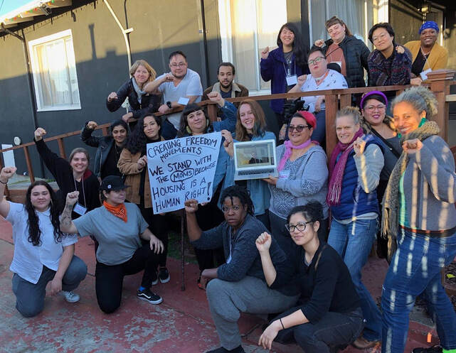 A group of CA group of 22 cohort members poses outside the Disability Justice Culture Club house with smiles and fists in the air to support the Moms4Housing movement. Two people hold a sign that reads: “Creating Freedom Movements - We Stand with the Moms because Housing Is a Human Right!”