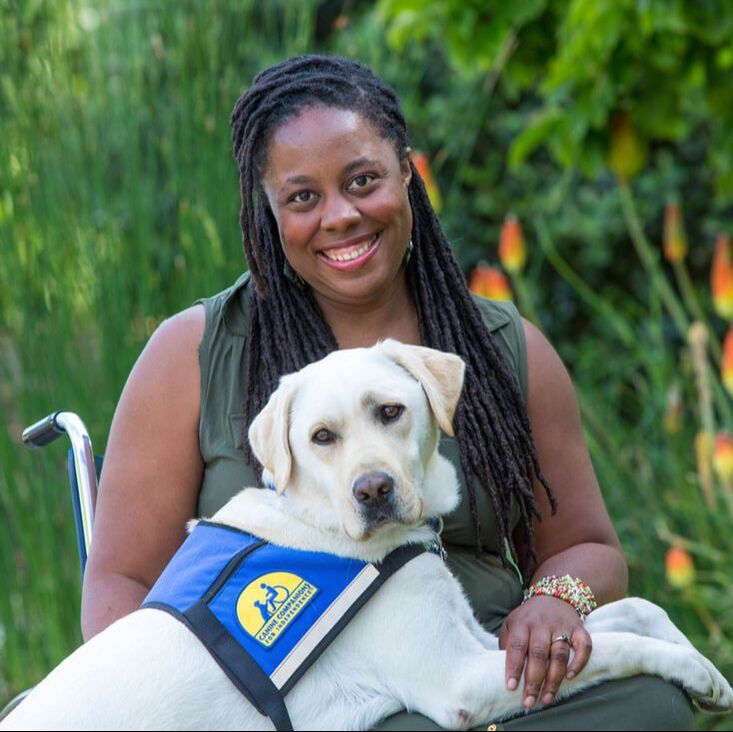 India Harville, African American female with long black locs, seated in her manual wheelchair wearing a long sleeveless green dress.  Her service dog, Nico, a blond Labrador Retriever, has his front paws on her lap.  He is wearing a blue and yellow service dog vest. They are outside with greenery behind them.